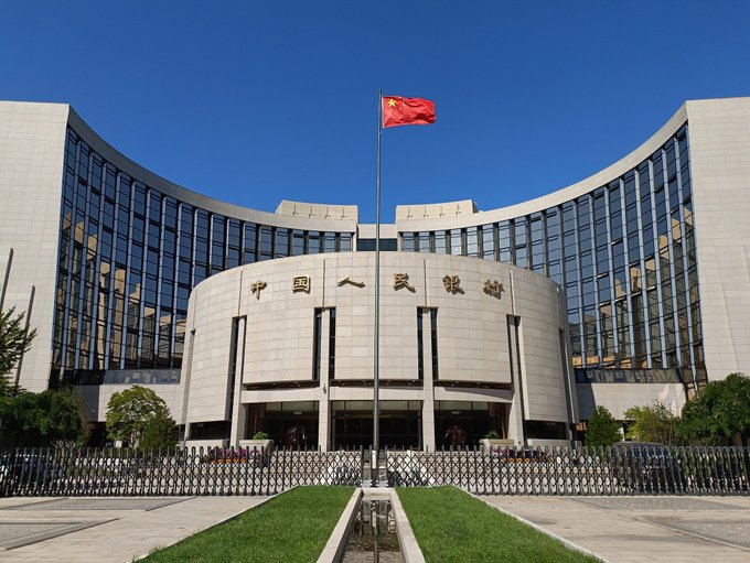 The People's Bank of China (中国人民银行) headquarters building with a Chinese flag flying in front under a clear blue sky.