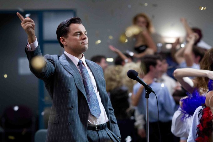 Leonardo DiCaprio as Jordan Belfort in the movie "The Wolf of Wall Street," wearing a pinstripe suit and pointing upward while standing at a microphone in front of cheering office workers with confetti falling.