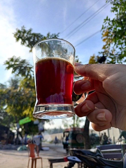 A hand holds up a glass mug of dark amber tea, likely chai, against an outdoor backdrop with trees, sky, and street activity.