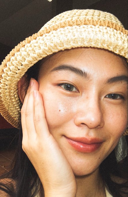 A close-up selfie of a young woman wearing a woven straw bucket hat, resting her face on her hand.