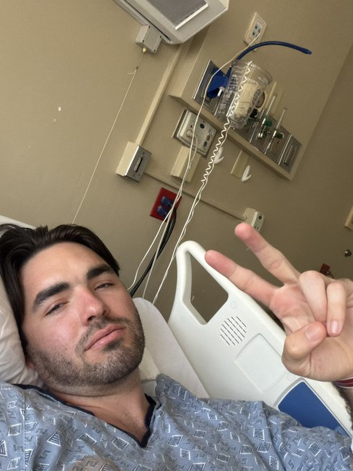 A man with dark hair and stubble lies in a hospital bed wearing a patterned gown, flashing a peace sign while medical equipment is visible on the wall behind him.