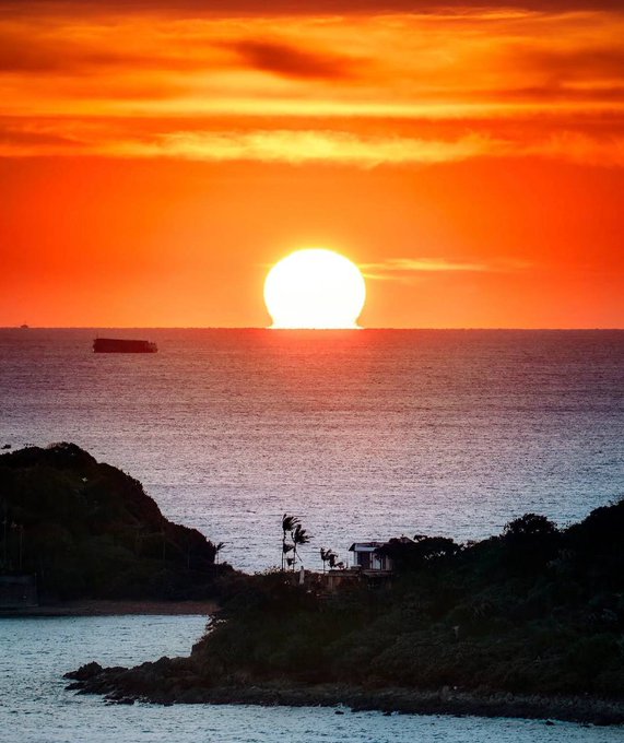 A vibrant sunset over the ocean with a large orange sun low on the horizon, a silhouette of a small boat, and a dark coastline in the foreground.
