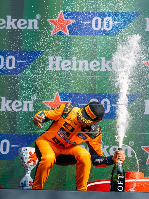 A racing driver in an orange suit celebrates on a podium by spraying a Moët champagne bottle, with Heineken branding and "0.0" displayed in the background.