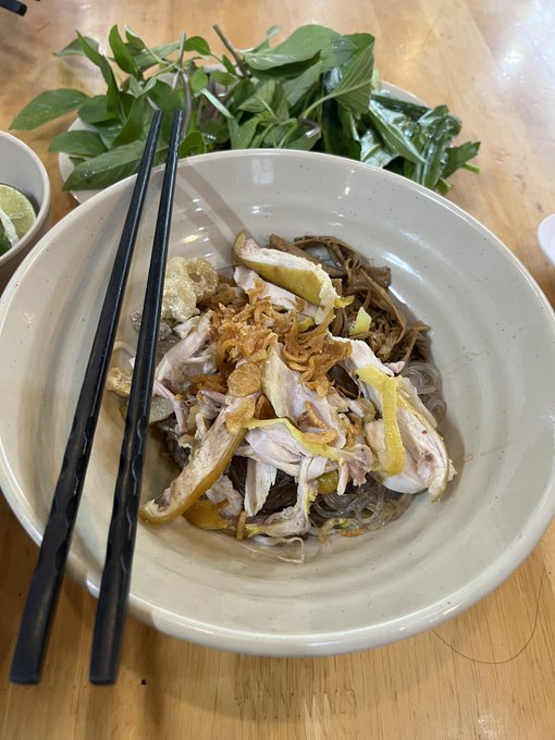 A close-up photo of a bowl of Vietnamese chicken and mushroom glass noodles (miến gà) topped with fried shallots, served with chopsticks and a side of fresh herbs and lime.