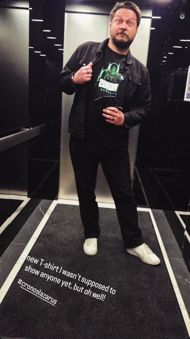 A bearded man stands in an elevator wearing a black jacket over a green "ZAMN!!!" graphic T-shirt, with overlaid text reading "new T-shirt! I wasn't supposed to show anyone yet, but oh well!! #cronoslazarus," teasing his upcoming role in #CronosNewDawn.