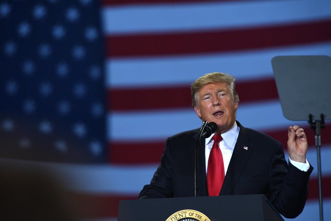 a person in a suit and red tie stands at a podium speaking in front of a large American flag backdrop.