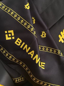 A black and yellow Binance-branded fabric, possibly a commemorative item for the exchange's 9th anniversary, featuring the "BINANCE" name and logo symbols. The caption relates to the image by celebrating the user's long-term loyalty to the platform, referencing the special anniversary gift shown.