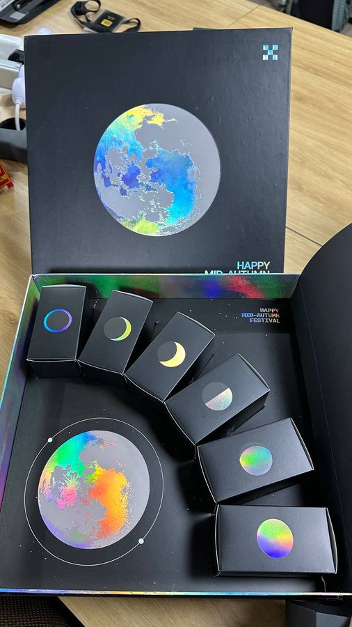 An open gift box labeled 'HAPPY MID-AUTUMN FESTIVAL' containing small black boxes with holographic moon phase designs and a larger image of the moon.