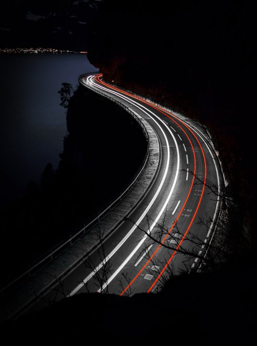 A long-exposure night photograph of a winding mountain road with bright white and red light trails from passing vehicles, with a dark lake and distant city lights visible in the background.