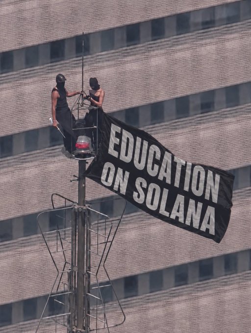 Two climbers atop a tower structure deploy a large black banner reading "EDUCATION ON SOLANA" against the backdrop of a building's facade, aligning with the post's description of GREED Academy students staging a bold stunt at the Empire State Building to advocate for Solana-based education.
