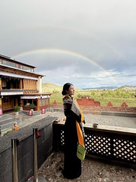 A woman in traditional Tibetan dress stands holding a baby, posing for a photo in front of a traditional building with a stunning rainbow arching across the cloudy sky behind the landscape.