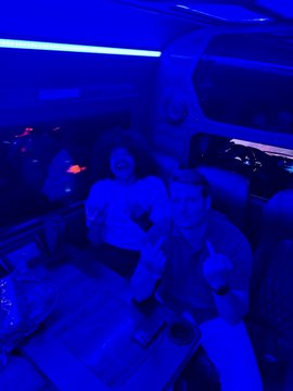 A dimly lit, blue-tinted photograph of three people sitting inside a vehicle, possibly a party bus or RV, with glowing lights and drinks on a table.