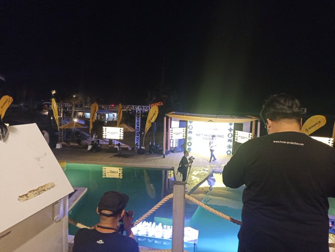 A nighttime scene shows a chess event by a pool with "NETWORKING AREA" signage, Binance logos visible, and a person in a black shirt with "www.how-pradelio.com" standing in the foreground.