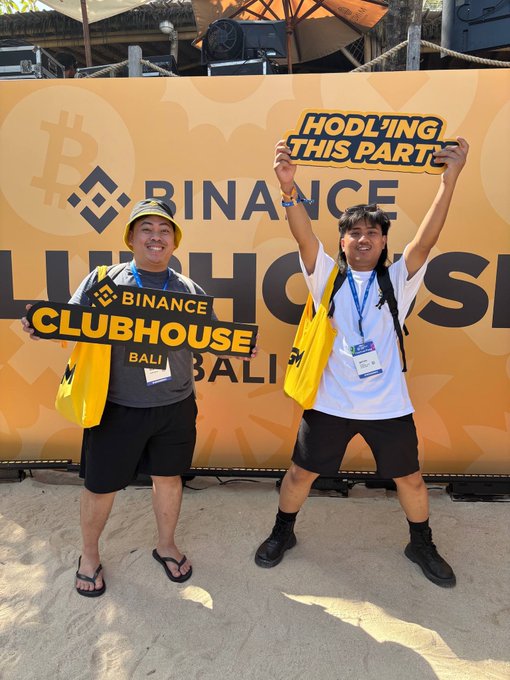 Two smiling people pose at a Binance Clubhouse Bali event, holding signs reading 'HODLING THIS PARTY' and 'BINANCE CLUBHOUSE BALI'.