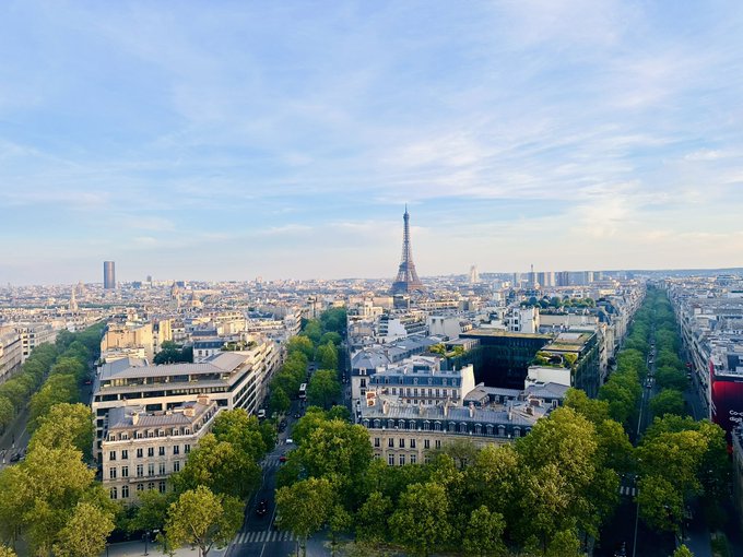 A scenic view of Paris from above, featuring the Eiffel Tower in the center, surrounded by classic Parisian architecture, tree-lined boulevards, and a partly cloudy sky.