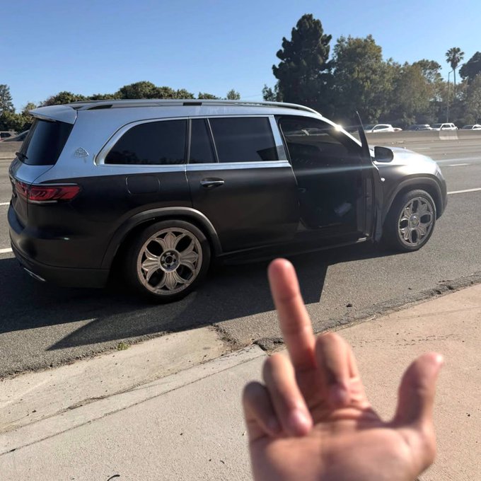 A matte black luxury SUV is parked on the side of a road with the driver's door open, while a hand in the foreground makes an obscene gesture toward the vehicle.