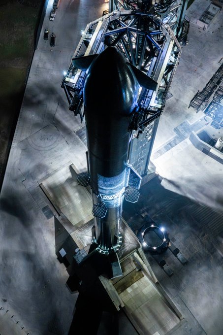 A top-down aerial view shows SpaceX's Starship rocket on its launch pad at night, surrounded by the launch tower's mechanical arms catching the vehicle. The image complements the post about owning shares in The Spaceship Company ($SPCX), visually representing the iconic rocket central to SpaceX's mission and the company's investment appeal.