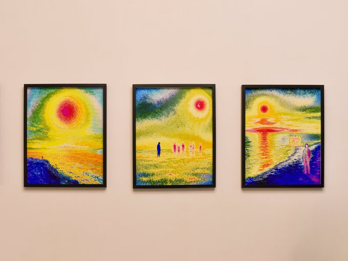 Three vibrant, expressionistic landscape paintings hang side by side in black frames, each featuring radiant sun motifs in red, yellow, and orange tones alongside small, ghostly human figures traversing dreamlike horizons. The works visually echo the exhibition's thematic tension—luminous celestial bodies glowing against vast landscapes mirror the "fragile boundary between visibility and existence," while the distant, barely-resolvable figures embody subjects caught between presence and disappearance.