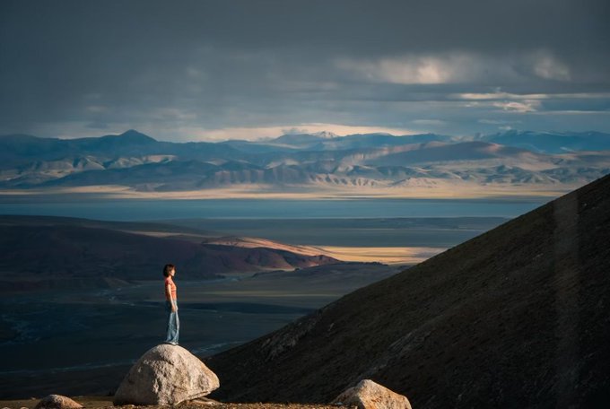 A scenic photograph of a person standing on a rock, looking out over a vast landscape with mountains, a lake, and a dramatic cloudy sky.