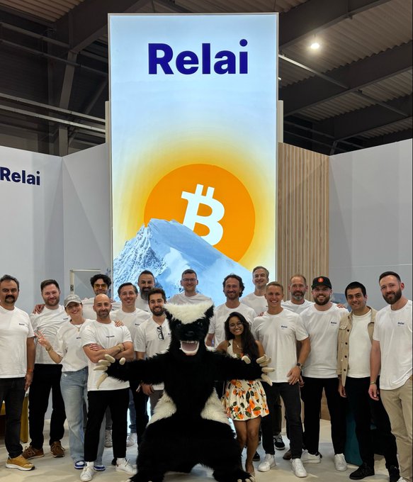 The image features a group of men posing for a photo with a mascot dressed as a black bear standing behind them. The mascot has a large orange sun on its back, which adds a fun and playful element to the scene. The people are wearing white t-shirts, suggesting that they might be part of an event or gathering related to cryptocurrency or blockchain technology. The post includes text such as "Relai" and "See you next year @BTCPrague", indicating that it is promoting an upcoming event or project in the world of cryptocurrencies.