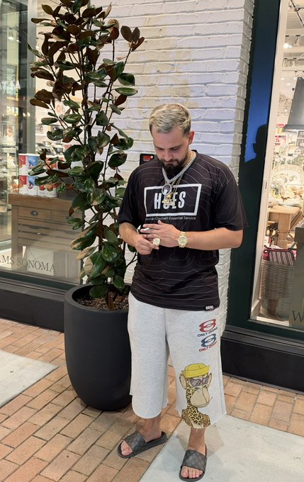 A man with gray hair stands on a tiled sidewalk looking at his phone, wearing a black t-shirt, gold chain and watches, gray shorts with logos, and slide sandals, with a potted plant and storefront visible behind him.