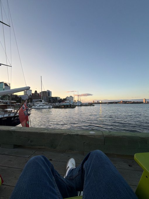 A first-person perspective photo shows a person's lap and legs as they relax on a bench overlooking a harbor at sunset, with a Canadian Red Ensign flag and boats visible in the background.