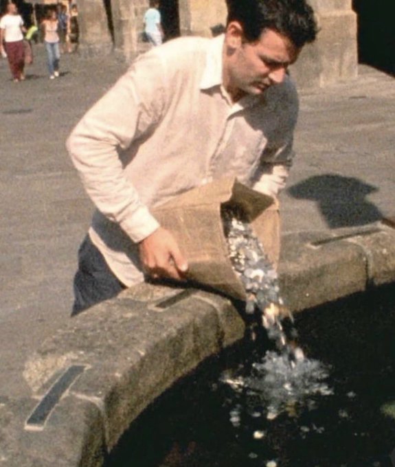 A man in a white shirt pours a sack of ice cubes into a stone fountain on a sunny city street.