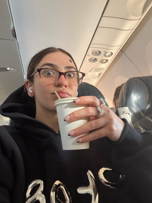 A woman wearing glasses and a black hoodie drinks from a white paper cup through a straw on an airplane, with a ring visible on her finger.
