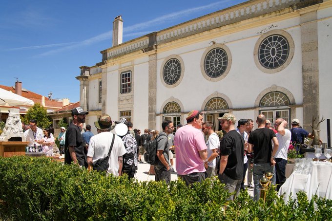 The image shows a large crowd of people gathered outside an old white building with circular windows. The people are standing around each other, engaged in conversation or observing something happening inside the building. Some individuals have backpacks, suggesting they might be tourists exploring the area. The scene appears to be set on a sunny day, as there is no indication of rain or overcast skies. The crowd's interest and interaction with their surroundings suggest that this location could be a popular spot for locals and visitors alike to enjoy a casual gathering or event.