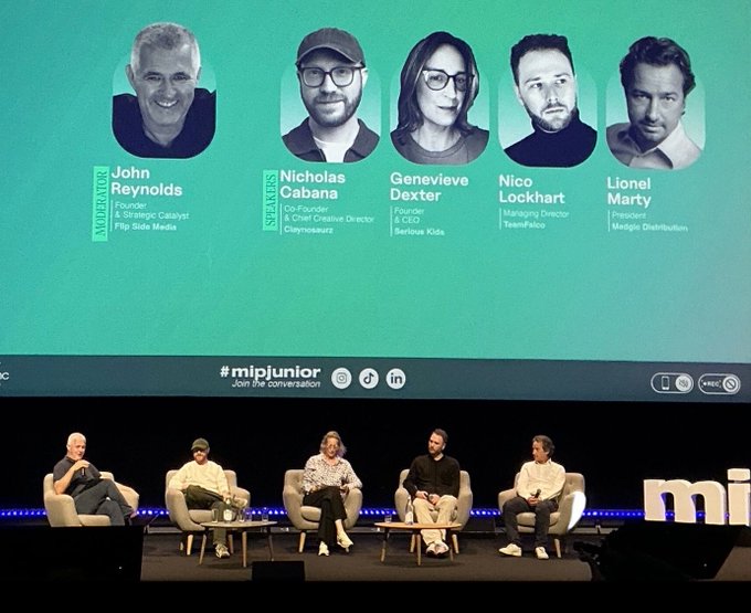 Five panelists—John Reynolds (Founder & Strategic Catalyst, Flip Side Media), Nicholas Cabana (Co-Founder & Chief Creative Director, Claynosaurz), Genevieve Dexter (Founder & CEO, Serious Kids), Nico Lockhart (Managing Director, TeamFellow), and Lionel Marty (President, Mediaprint/Budding)—sit on stage at the MIPJUNIOR conference in Cannes, with their names and titles displayed above them on a teal backdrop featuring the #mipjunior hashtag. The image illustrates the panel discussion mentioned in the post, where Claynosaurz (represented by Nicholas Cabana) joined other industry leaders to explore new approaches to funding and building original kids' and family entertainment IP.