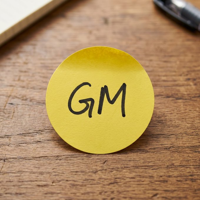 A yellow circular sticky note with 'GM' handwritten in black marker, sitting on a wooden surface next to a pen.