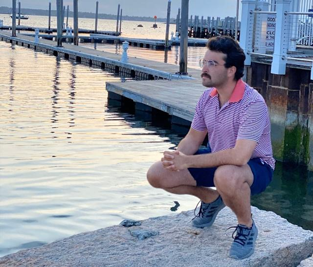 A man in a striped polo shirt squats on a rock by a marina with wooden docks and a sign listing dog rules, gazing contemplatively across the water—evoking the somber, reflective mood tied to the post about the collapsed GPT models SOL, TERRA, and LUNA under the 5.6 umbrella.