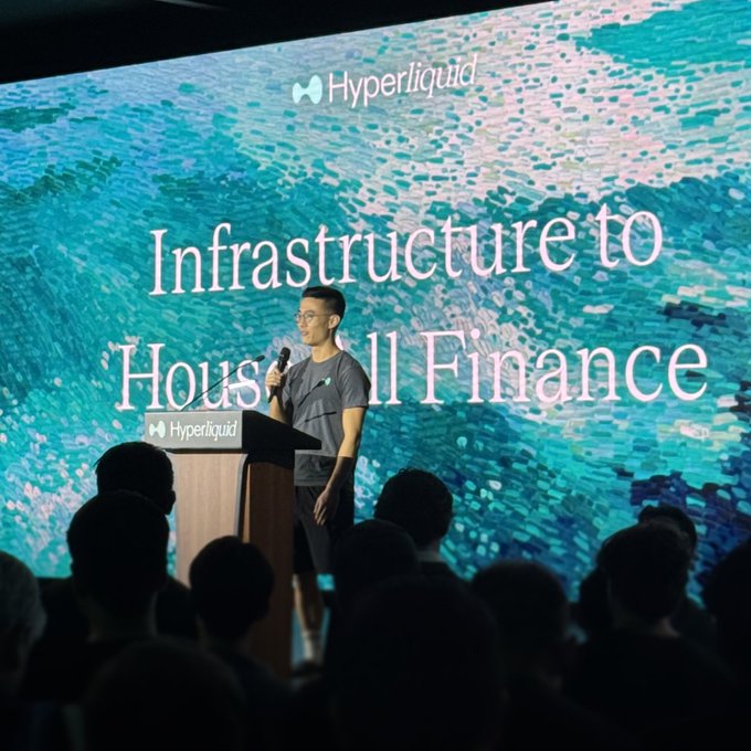 A man speaks at a podium branded with the Hyperliquid logo in front of a large screen displaying 'Infrastructure to Household Finance' against a shimmering teal background.