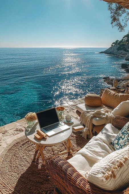 A serene Mediterranean coastal scene featuring a laptop on a small table set up on a cliffside terrace with cushions, overlooking a sparkling blue sea under bright sunlight.