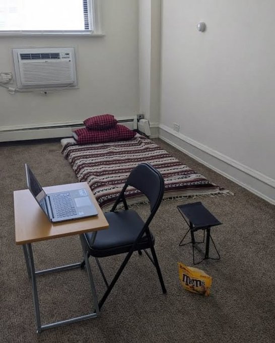 A makeshift workspace features a mattress on the floor beside a simple desk holding a laptop, paired with a folding chair and an even smaller foldable stool, with a bag of "m&m's" candy sitting on the carpet. The scene humorously reflects the tweet's "Number of employees: 1" caption, depicting the sparse, humble "HQ/office" of a tiny startup or solo developer operation.
