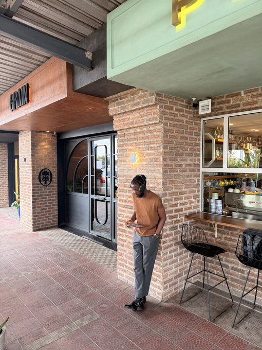 A man wearing headphones and a brown shirt leans against a brick column outside a café or bar named 'BRÜNT', looking down at his phone, with a window display of bottles and stools visible behind him.