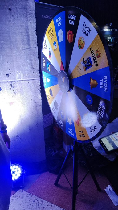 A branded prize wheel from 'noOnO' featuring various rewards like a dog fan, wool tee, luck cap, and Bybit tee is set up at an event with blue lighting.