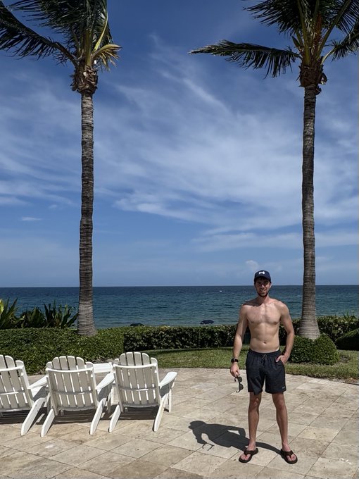 A shirtless man in a black cap and swim shorts stands on a patio between two palm trees overlooking the ocean in Boca Raton, appearing to casually enjoy the beachside view. The caption's contrast between his relaxed, sunny surroundings and the mention of "FOMO long on XRP" humorously highlights the tension between vacation bliss and crypto market anxiety.