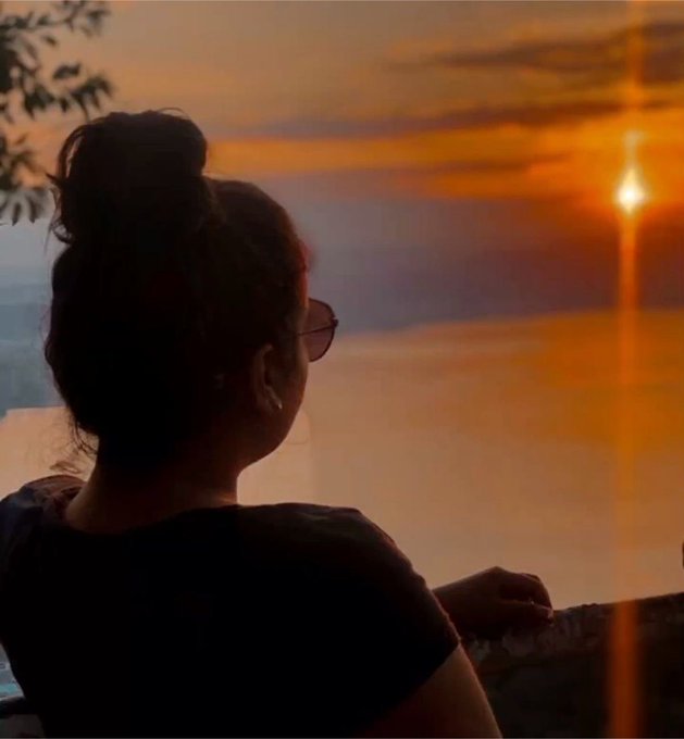 Silhouette of a woman with her hair in a bun, wearing sunglasses and hoop earrings, watching a bright orange sunset over the ocean.