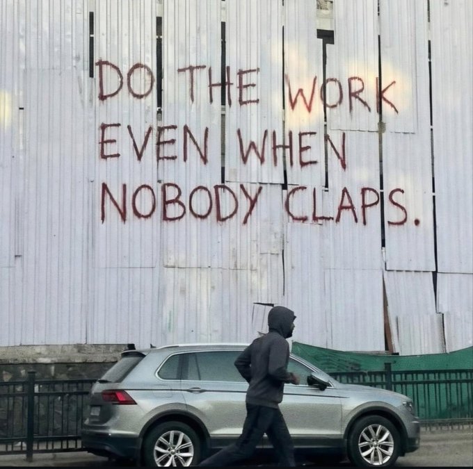 A person in a dark hoodie walking past a silver SUV in front of a corrugated metal wall with red spray-painted graffiti that reads 'DO THE WORK EVEN WHEN NOBODY CLAPS.'