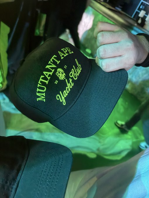 A hand holds a black cap with neon green text reading 'MUTANT APE Yacht Club' alongside a skull logo, set against a green-lit background.
