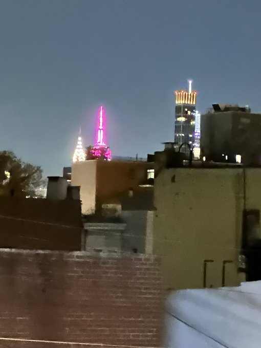 A nighttime cityscape showing rooftops in the foreground with brightly lit skyscrapers in the distance, including one with a purple-lit spire.