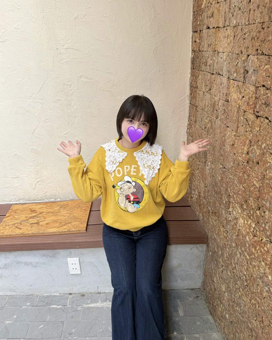 A young woman wearing a yellow sweater featuring a Popeye graphic and a white lace collar stands with her arms raised; her face is obscured by a purple heart emoji.