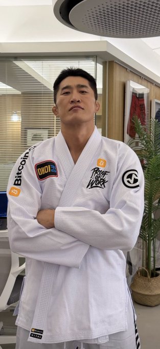 A confident man in a white Brazilian Jiu-Jitsu gi stands with arms crossed, his uniform prominently displaying "Bitcoin.com" and "Proof of Work" patches, aligning with the post's playful jab about the recently launched physical Bitcoin.com martial arts gear.