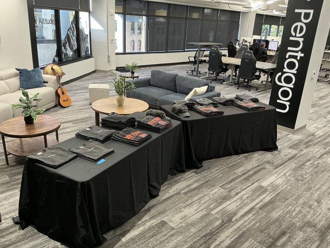 An office lounge area named 'Pentagon' with tables displaying folded branded t-shirts, modern furniture including a gray sofa and armchairs, a guitar, and workspace with chairs and monitors in the background.