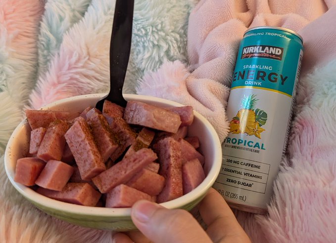 A hand holding a bowl of cubed Spam sprinkled with seasoning, accompanied by a can of Kirkland Signature Sparkling Tropical Energy Drink (200 mg caffeine, zero sugar), set against a fluffy blanket background.