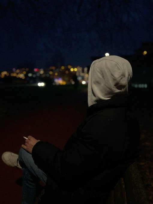 A person wearing a white hoodie and black jacket sits on a bench at night, holding a cigarette, with blurred city lights visible in the dark background.