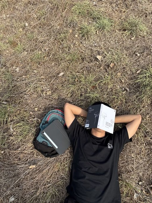 A person lying on dry grass with a book covering their face, wearing a black t-shirt, with a backpack and tablet resting beside them.
