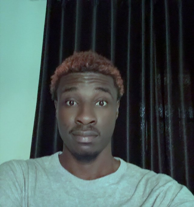 A selfie of a young man with reddish-brown hair wearing a gray t-shirt, photographed against a dark curtain background.