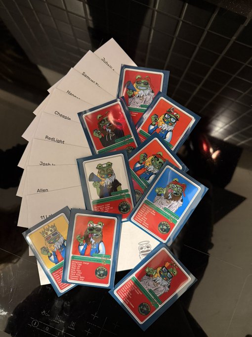 A collection of frog-themed character trading cards with various attributes spread out on a dark surface, alongside several envelopes labeled with names like 'Cheesie', 'RedLight', 'Josh', 'Allen', and 'Hammer'.